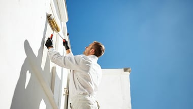 professional painter painting exterior wall of house with paint roller, blue sky